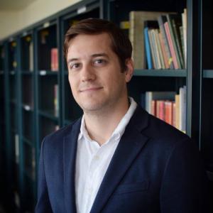 Photo of Professor Mazanec in a blue blazer and white shirt, in front of bookshelves, looking scholarly